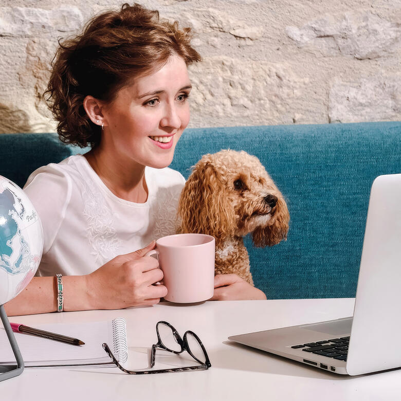 Smiling woman sitting at laptop with a small poodle beside her.